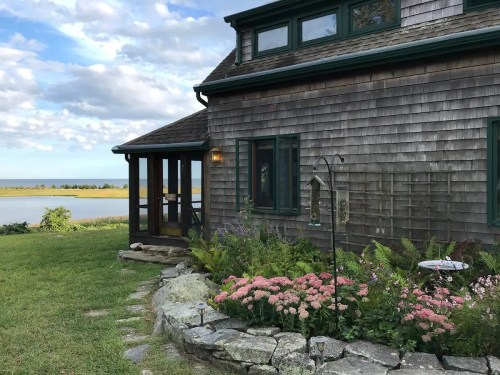 A charming wooden house with a garden of pink flowers, overlooking a serene body of water under a cloudy sky.