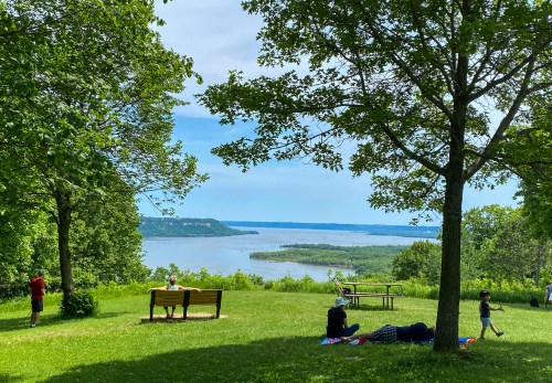 A scenic view of a river surrounded by greenery, with people enjoying a picnic under trees on a sunny day.
