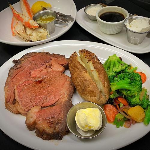 A plate featuring prime rib, baked potato with butter, steamed broccoli, and carrots, with crab legs and dipping sauces in the background.