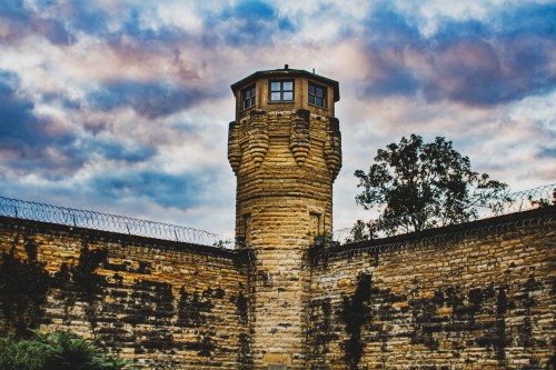 A stone prison tower with a watchtower, surrounded by a high wall and a cloudy sky in the background.