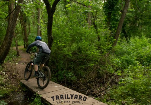 A mountain biker rides over a wooden bridge in a lush, green forest. The scene is vibrant and full of foliage.
