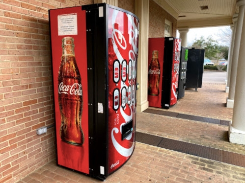 Two Coca-Cola vending machines are positioned outside a building, with a brick walkway in front.