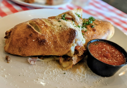 A golden-brown calzone filled with meat and cheese, served with a side of marinara sauce and sprinkled with parsley.