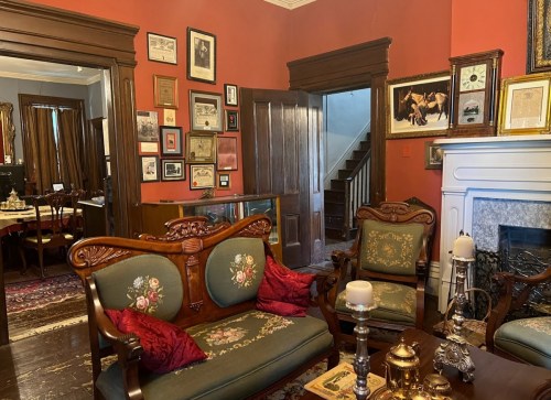 A vintage living room with ornate furniture, framed pictures on the walls, and a staircase in the background.