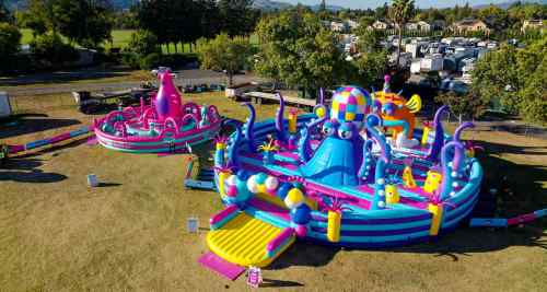 Colorful inflatable water park with slides and playful characters, set in a grassy area with trees in the background.