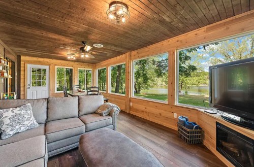 Cozy sunroom with wooden walls, large windows overlooking a lake, a sofa, and a TV. Bright and inviting atmosphere.