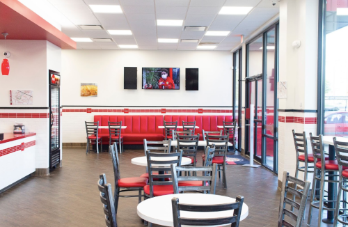 Bright, modern restaurant interior with red accents, tables, chairs, and a TV displaying a person.