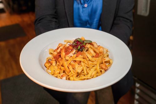 A person in a suit holds a plate of fettuccine pasta topped with seafood and garnished with herbs.
