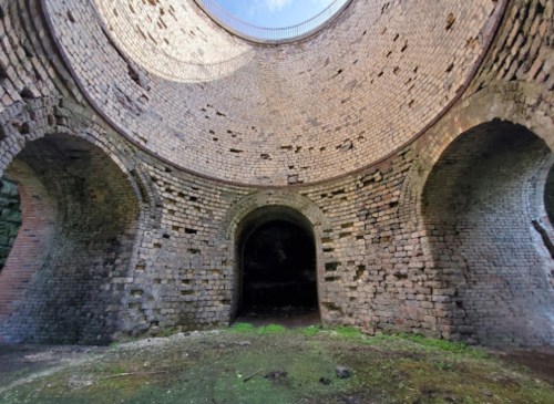 Interior view of a large, circular brick structure with arched openings and a partially open roof, overgrown with moss.