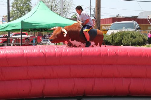 A child rides a mechanical bull on a red inflatable surface at an outdoor event, with tents in the background.