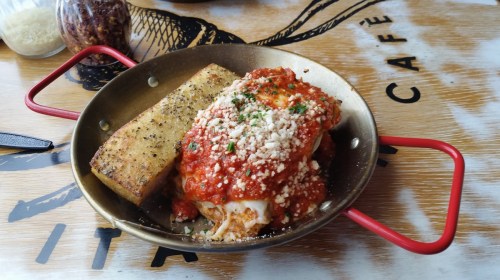 A skillet with baked pasta topped with marinara sauce, cheese, and a side of toasted bread on a wooden table.