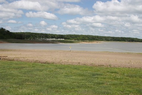 A serene lakeside view with a grassy foreground, cloudy sky, and a person standing near the water's edge.