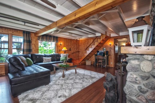 Cozy living room with a black sectional sofa, wooden beams, a stone fireplace, and a staircase leading to an upper level.