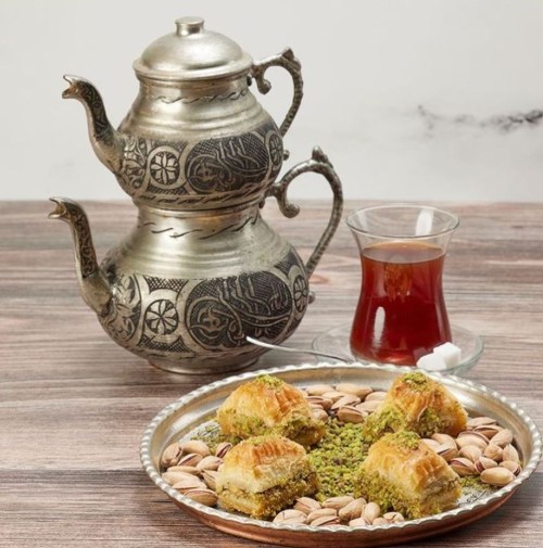 A silver teapot set with a glass of tea and a plate of baklava topped with pistachios and almonds.