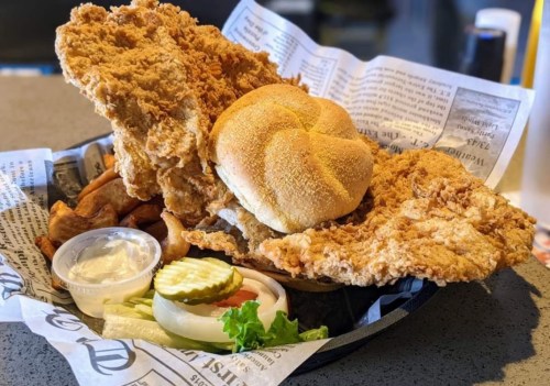 A basket of crispy fried chicken, a sandwich bun, pickles, lettuce, and dipping sauce on a bed of newspaper.