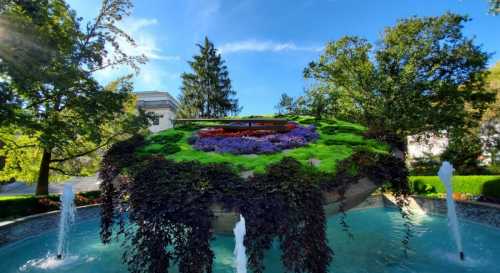 A vibrant circular garden atop a fountain, surrounded by lush greenery and colorful flowers under a clear blue sky.