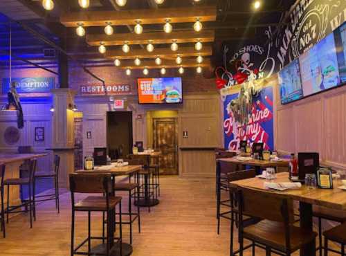 Interior of a restaurant featuring wooden tables, bar stools, and decorative lighting, with screens and a rustic vibe.