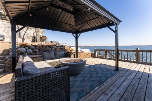 A cozy outdoor gazebo with seating and a fire pit, overlooking a serene lake on a clear day.
