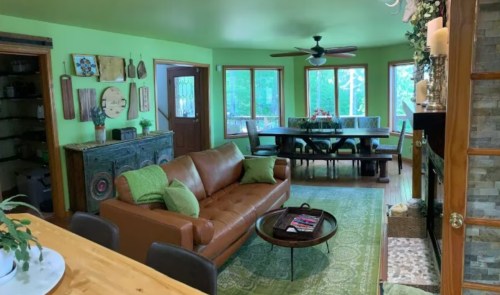 Cozy living room with green walls, a brown leather sofa, dining area, and large windows overlooking greenery.