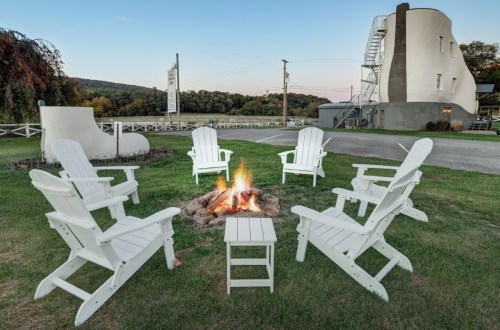 A cozy fire pit surrounded by white Adirondack chairs, with a scenic backdrop of hills and a unique building.