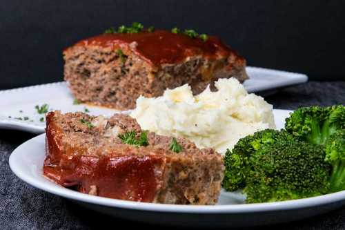 A plate featuring sliced meatloaf with ketchup glaze, mashed potatoes, and steamed broccoli on a dark background.