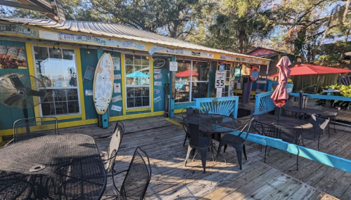 Colorful outdoor dining area with tables, umbrellas, and a surfboard on a vibrant building surrounded by trees.