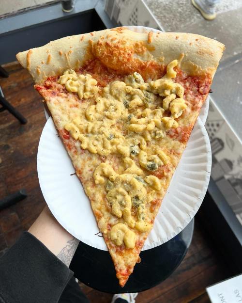 A large slice of pizza topped with macaroni and cheese, held above a table with a window view in the background.
