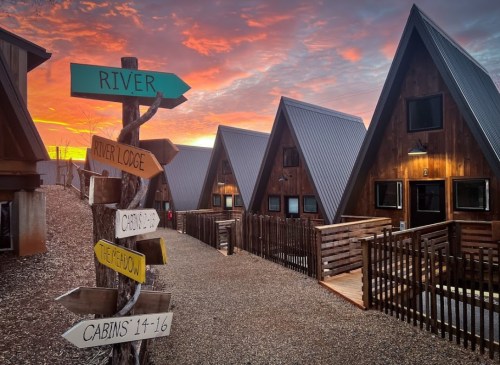 A scenic view of A-frame cabins at sunset, with a signpost pointing to various locations like "River" and "Cabins 1-16."