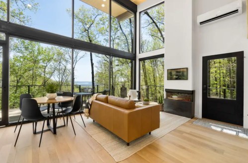 Modern living room with large windows, a brown sofa, dining table, and views of lush greenery outside.