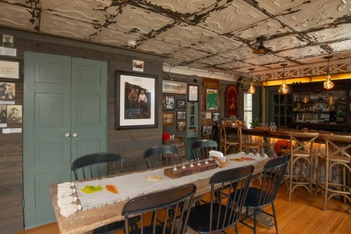 A cozy dining area with a long table, wooden chairs, and a bar, featuring vintage decor and a tin ceiling.