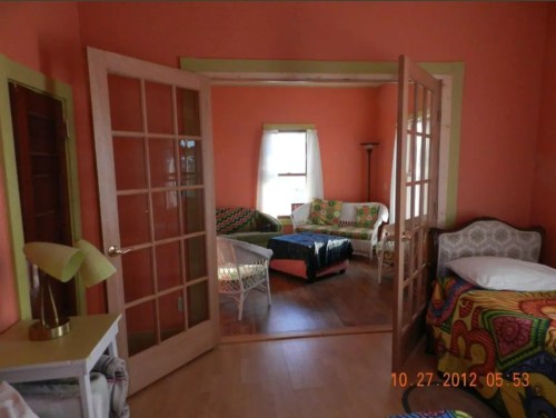 A cozy room with orange walls, featuring a seating area with colorful quilts and a lamp, viewed through double doors.