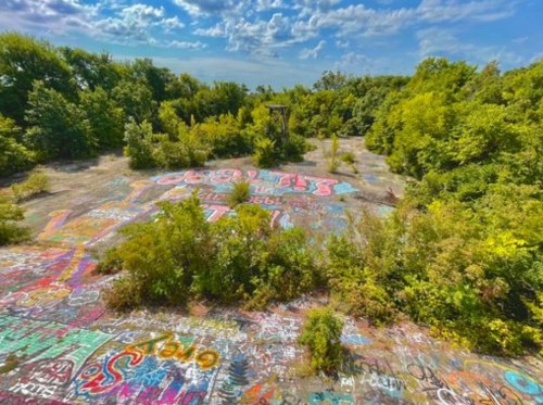 A vibrant, overgrown area covered in colorful graffiti, surrounded by lush greenery and a blue sky with clouds.
