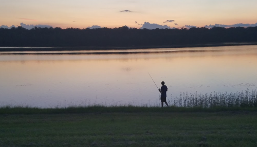 A person fishing by a calm lake at sunset, with a colorful sky reflecting on the water.