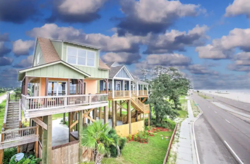 A colorful beach house on stilts with a scenic view, surrounded by greenery and a road leading to the beach.