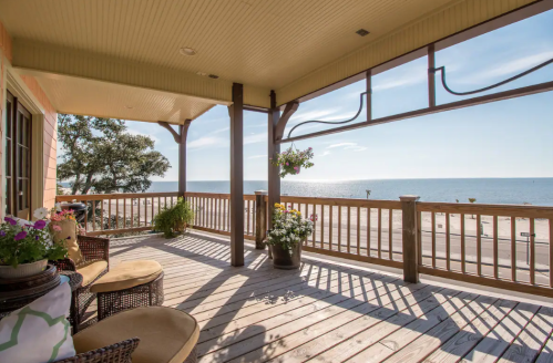 A sunny porch with wooden railings overlooks a calm sea, featuring potted plants and comfortable seating.