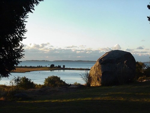 A large boulder sits near a calm waterway, with trees framing the view and clouds in the sky at sunset.