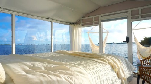 Cozy bedroom with a bed, large windows, and a view of the water, featuring hanging chairs and soft natural light.