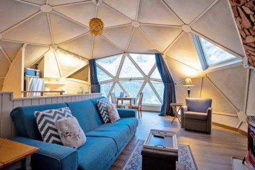 Cozy interior of a geodesic dome with a blue sofa, dining area, and large windows showcasing a scenic view.