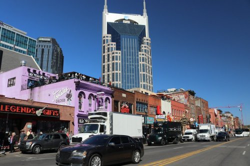 A bustling street scene in Nashville with colorful buildings, shops, and a tall skyscraper in the background under a clear blue sky.
