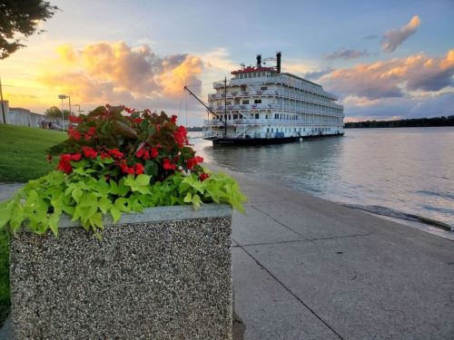 A riverboat docked near a flower planter at sunset, with colorful clouds reflecting on the water.