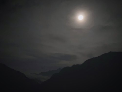 A full moon illuminates a dark, cloudy sky over shadowy mountain silhouettes.