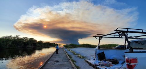 A person stands on a wooden dock by a river, watching a large plume of smoke rising into the sky at sunset.