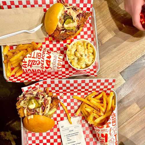 Two trays of food featuring fried chicken sandwiches, fries, and macaroni and cheese on a checkered paper background.