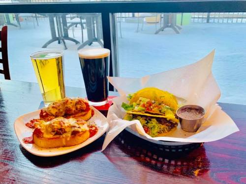 A plate with cheesy bread and a taco, accompanied by two glasses of beer on a table by a window.