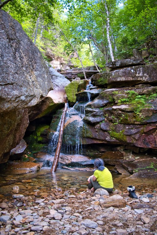 A person sits by a small waterfall surrounded by rocks and greenery, enjoying the serene natural setting.
