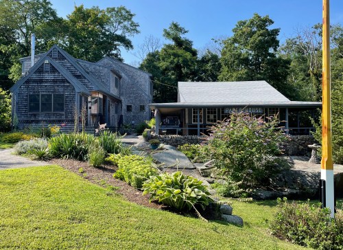 A charming two-story house with a porch, surrounded by lush greenery and colorful flowers on a sunny day.