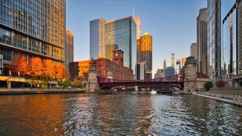 Cityscape of Chicago at sunset, featuring skyscrapers, a bridge over the river, and autumn trees along the waterfront.