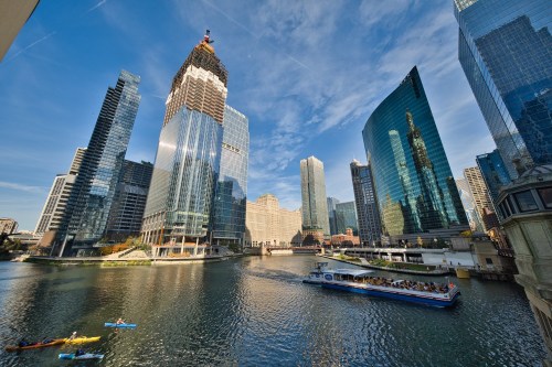 A vibrant city skyline with modern skyscrapers reflecting in the river, and boats navigating the water under a clear blue sky.