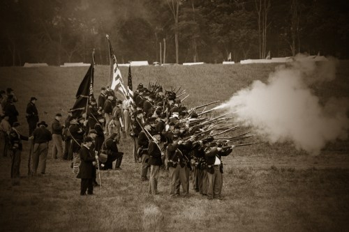 A sepia-toned image of soldiers in uniform firing rifles during a reenactment, with flags waving in the background.