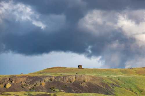 A dramatic sky looms over a green hillside with rocky outcrops, creating a striking natural landscape.